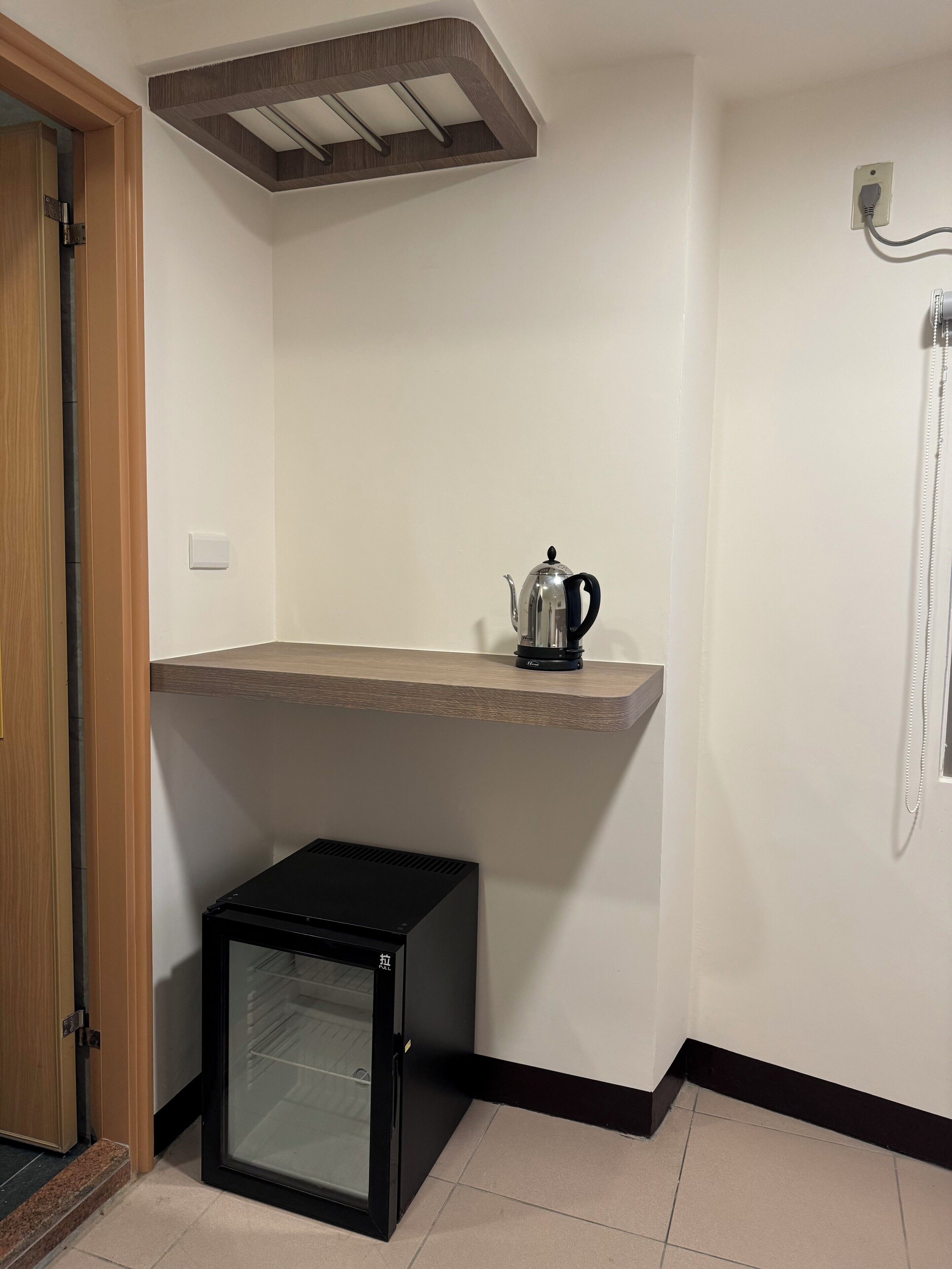A compact room with a kitchenette, a small refrigerator, a wooden shelf, and a built-in shower.