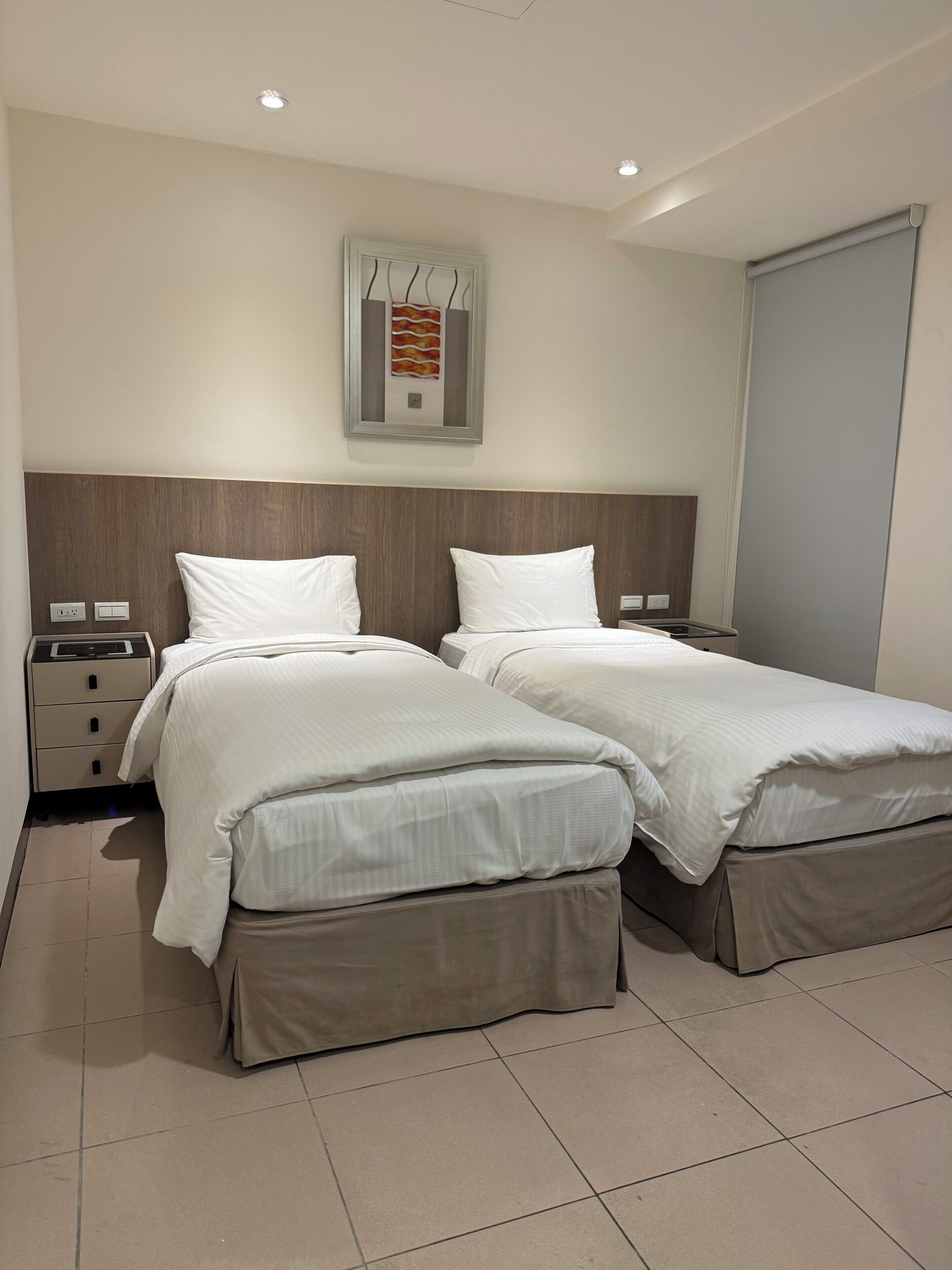 A hotel room with two beds, a wooden headboard, a wall-mounted artwork, a bedside table with drawers, and a large window.