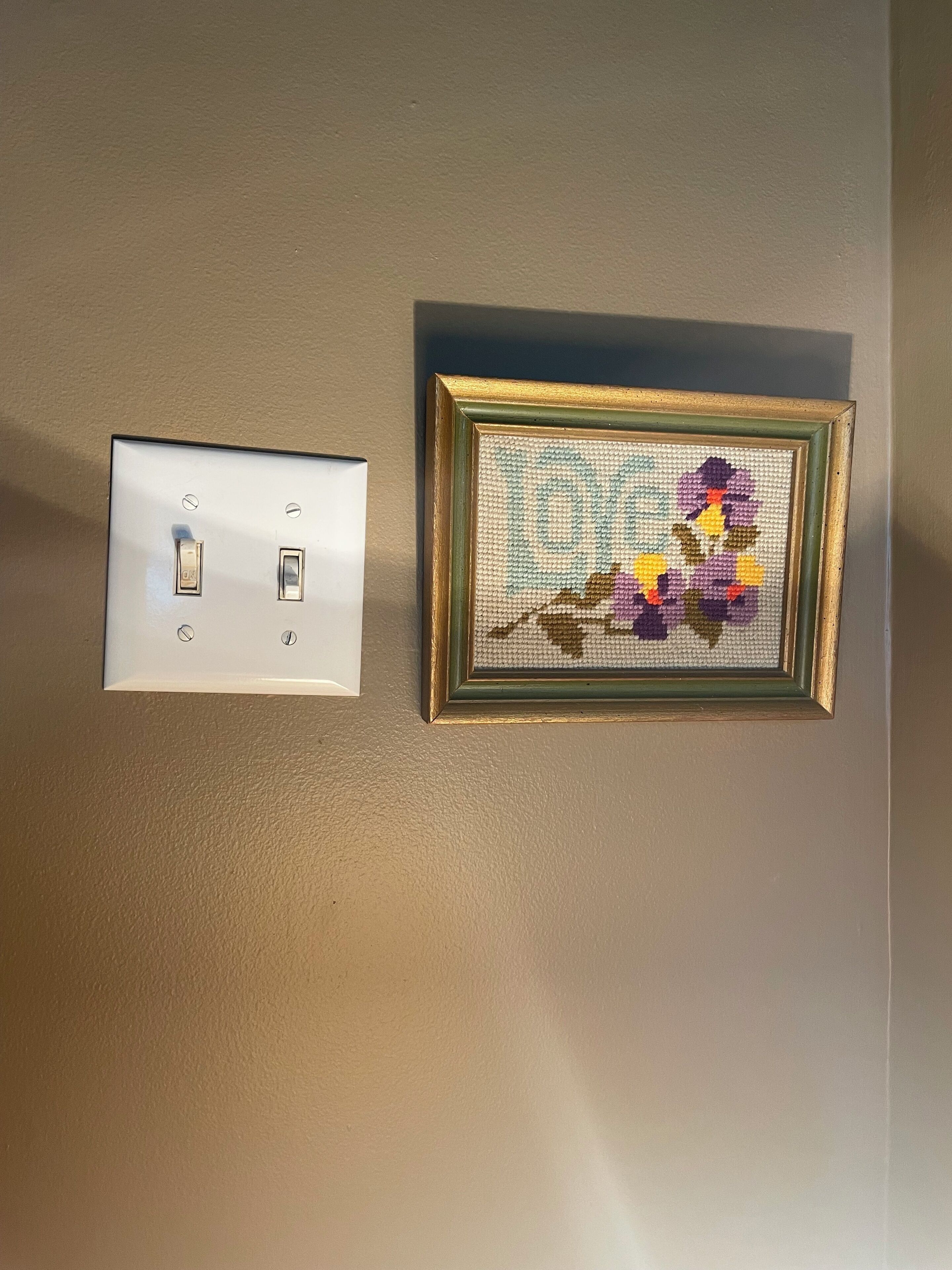 Een lichtschakelaar bevestigd aan de muur en een geborduurd kunstwerk in een lijst.