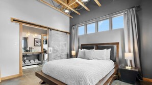 A modern bedroom with a large bed, a wooden headboard, and a view of a bathroom through an open sliding door.