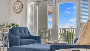 A living room with a blue armchair, a sofa, and a dining table with chairs. There is a large window with a view of the outside. - Spring Discounts! Pool & Hot tub.1st Fl. (Miramar Beach)