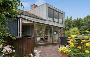 A modern two-story house with large glass windows, a wooden deck, and a garden with flowers. - 2 bedroom nice home in Juelsminde (Juelsminde)