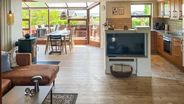 A spacious living area with a large TV, wooden flooring, a cozy seating area, and a kitchen with wooden cabinets and a large window. - 2 bedroom nice home in Juelsminde (Juelsminde)