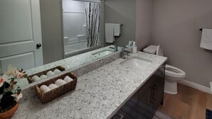 A modern bathroom with a large mirror, a sink, and a toilet. There are towels hanging on the wall and a basket of toiletries on the counter. - Separate Entrance Basement Suite (Winnipeg)