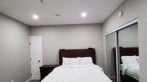 A bedroom with a bed, a door, a nightstand, a mirror, and a ceiling light. - Separate Entrance Basement Suite (Winnipeg)