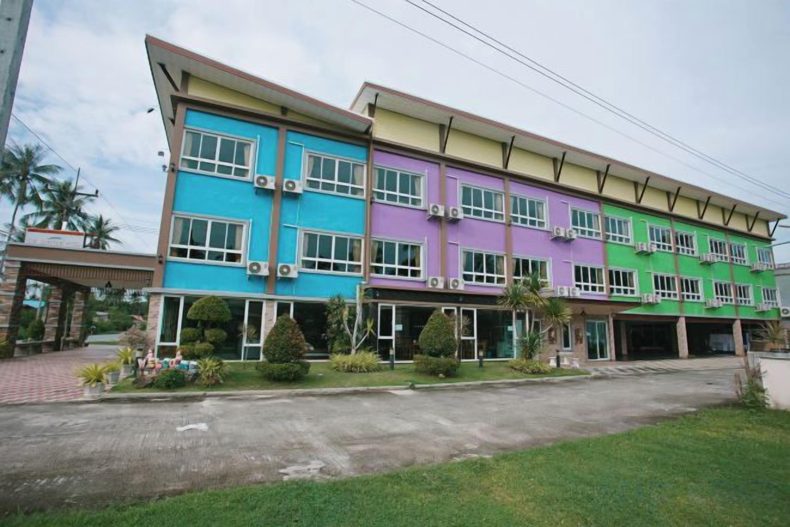 A multi-story building with a flat roof, multiple balconies, and a paved entrance area with a small garden.
