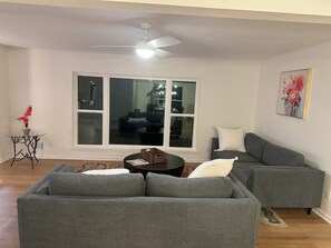 Ein Wohnzimmer mit grauem Ecksofa, einem kleinen runden Tisch und einem großen Fenster mit Blick ins Freie.