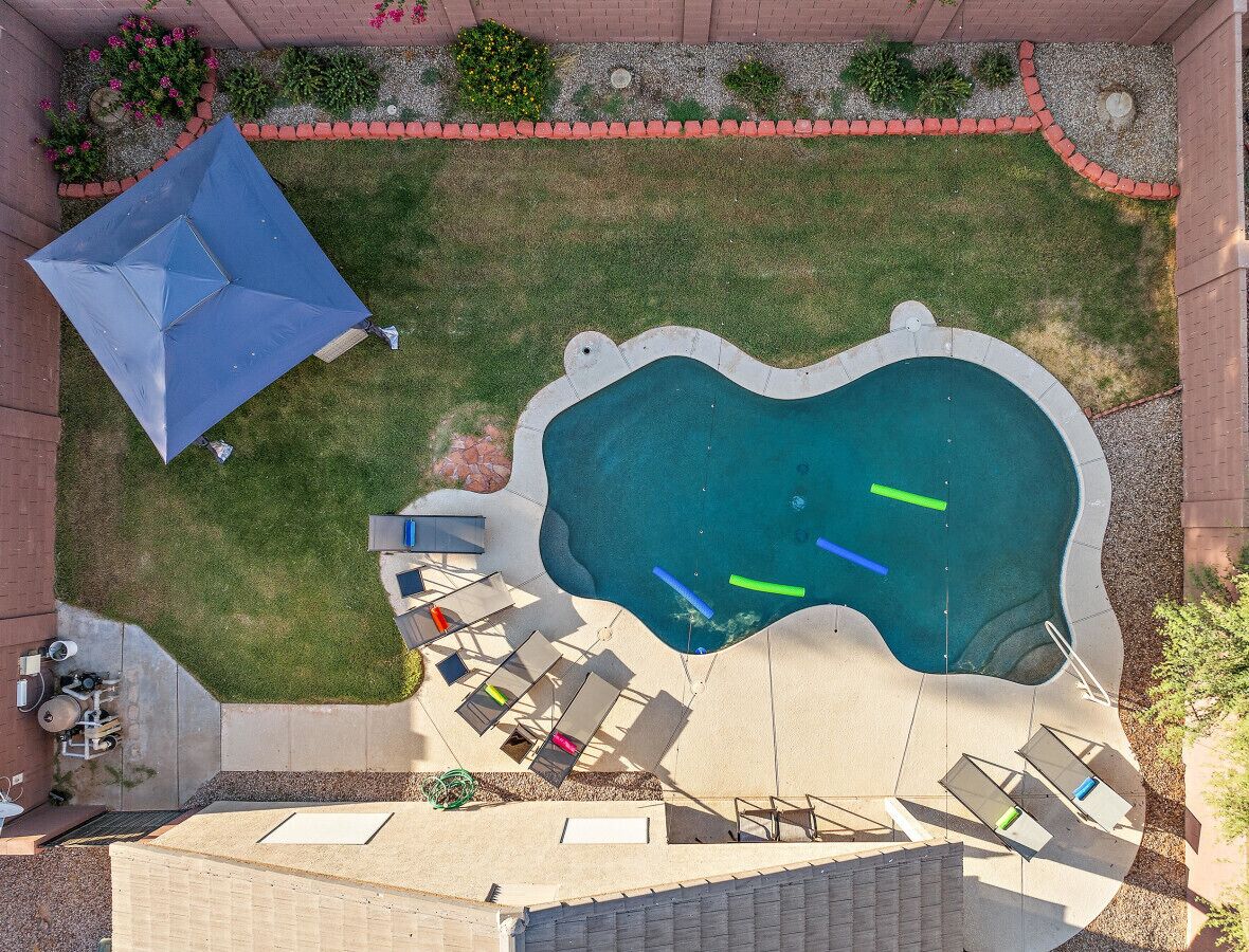 Ein Pool im Freien mit wellenförmigem Rand, umgeben von einer Betonplattform. Liegestühle und ein blauer Sonnenschirm sind vorhanden.