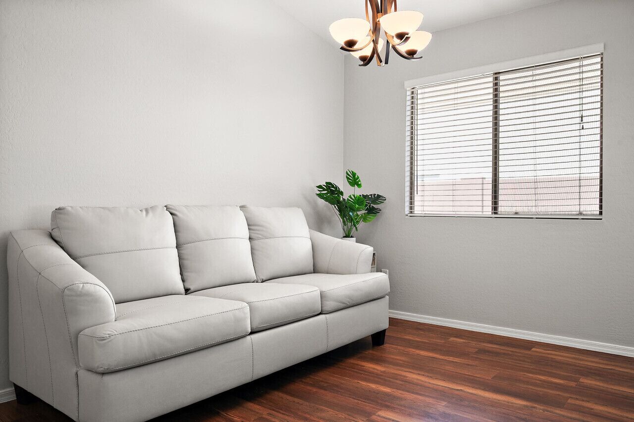 Ein Wohnzimmer mit einem hellen Sofa, einer Topfpflanze und einem Fenster mit Jalousien.