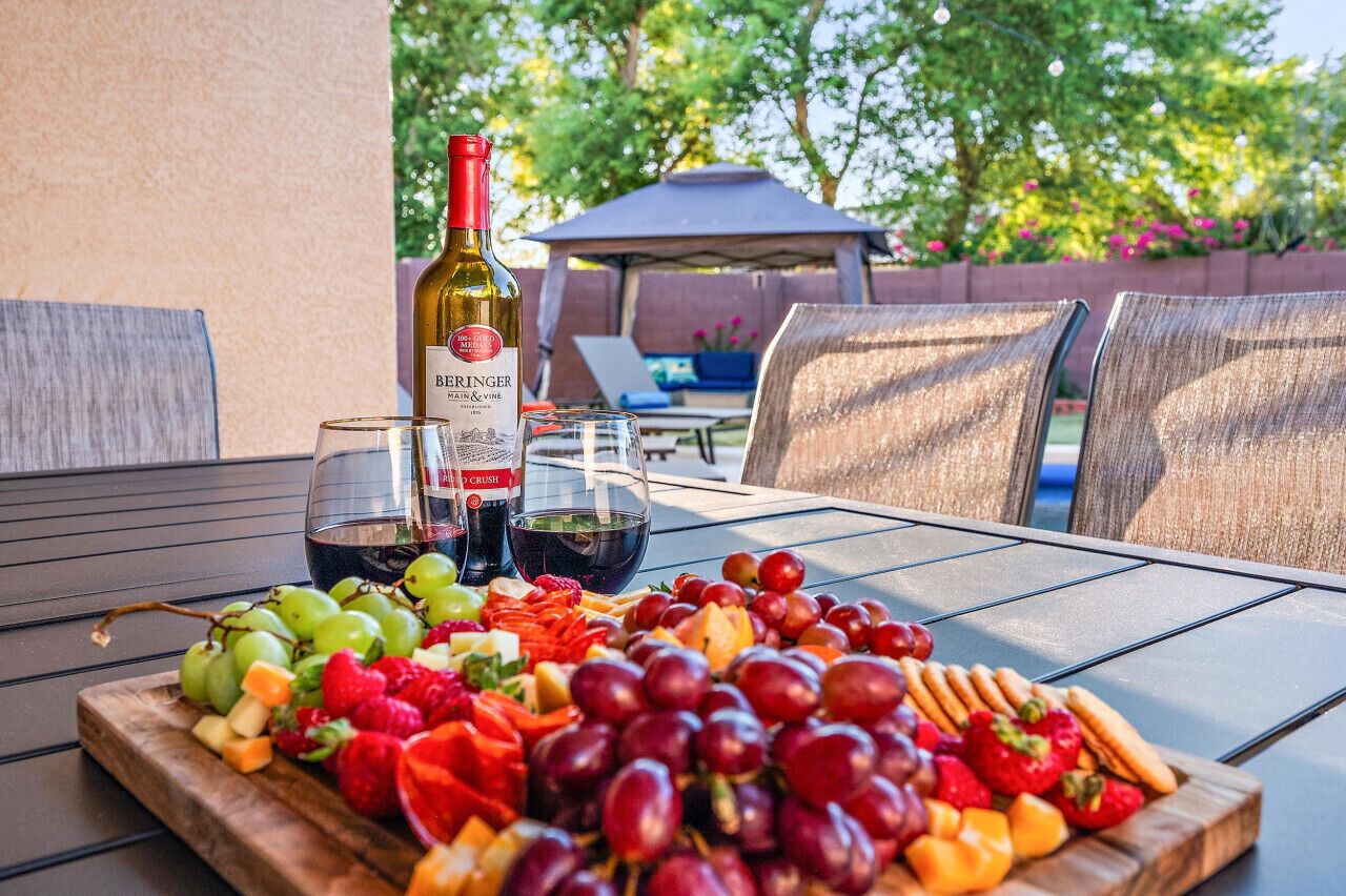 A table set outdoors with a bottle of wine, glasses, and a variety of fruits and snacks.
