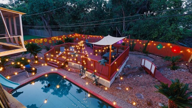 Un patio trasero con piscina, terraza de madera y luces colgantes.