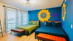 Una habitación con dos camas, una ventana con cortinas, una lámpara en el techo y un cuadro de girasoles en la pared.