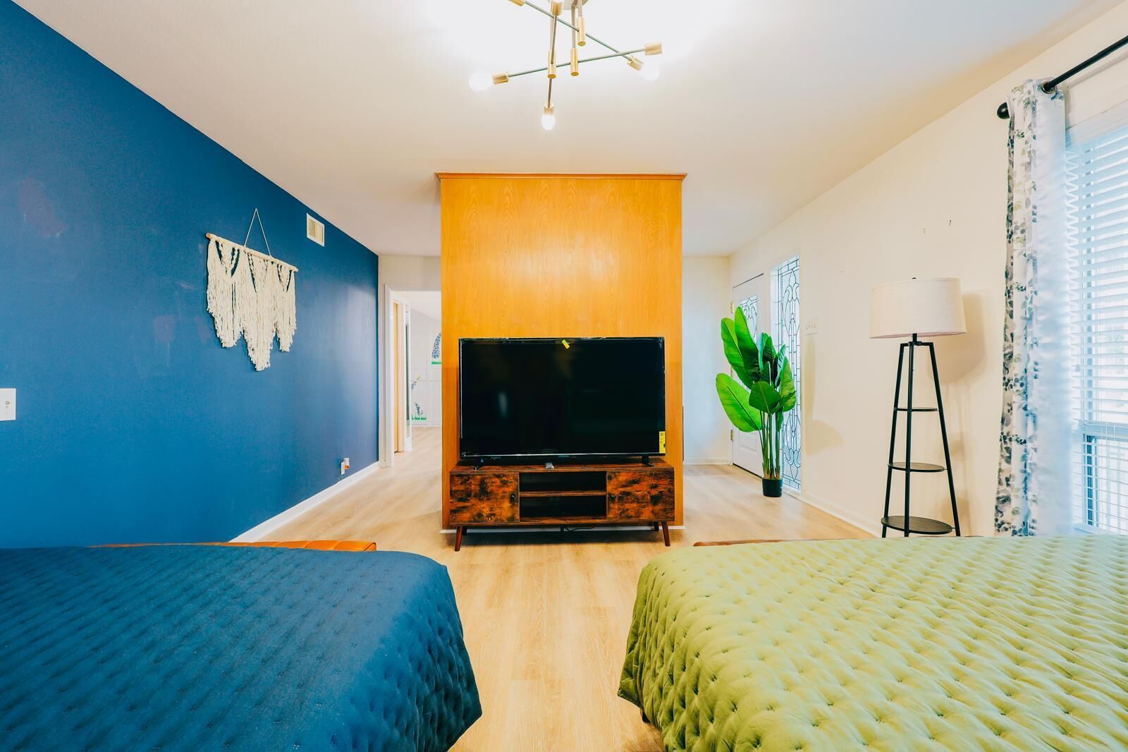 A hotel room with a large flat-screen TV, two beds, a wooden entertainment unit, a potted plant, and a floor lamp.
