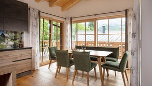 Un comedor con una mesa de madera y cuatro sillas, ventanas amplias con vistas a una terraza de madera y montañas, y una cocina con encimera oscura y armarios de madera.