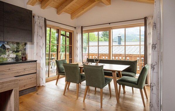 Un coin repas avec une table en bois et quatre chaises, de grandes fenêtres offrant une vue sur une terrasse en bois et les montagnes, et une cuisine dotée d’un plan de travail foncé et de meubles en bois.