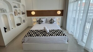 Une chambre moderne avec un grand lit, deux lampes suspendues, une tête de lit en bois et une vue sur l’extérieur à travers des rideaux transparents.