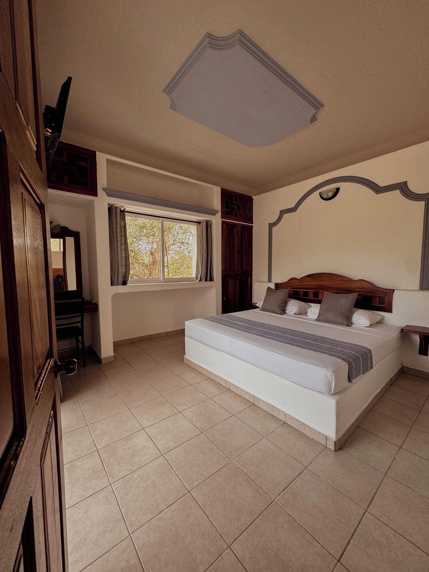 A bedroom with a large bed, a window with curtains, a wooden headboard, and a tiled floor.