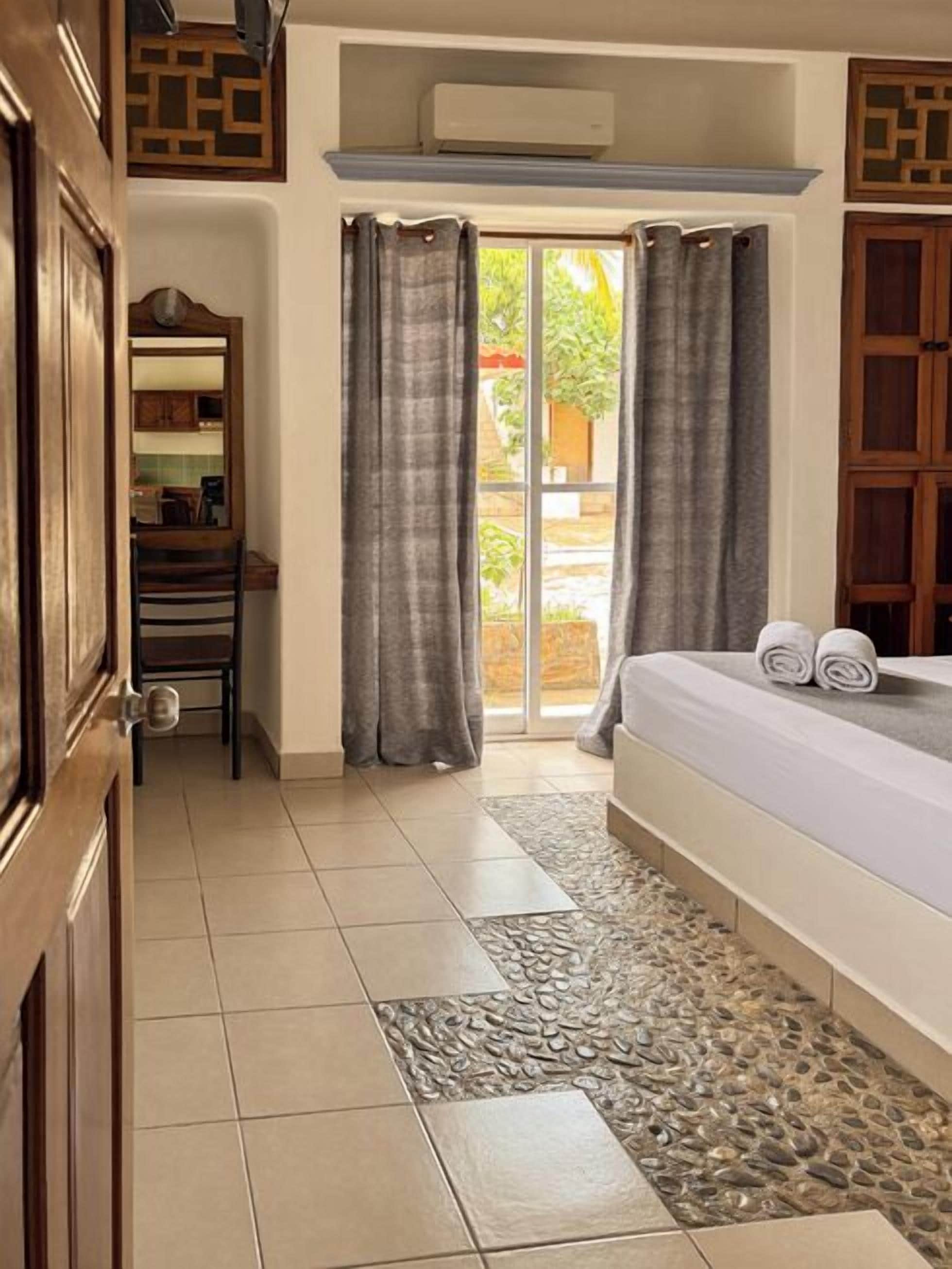 A hotel room with a bed, a small desk, a chair, and a view through a glass door to an outdoor area.