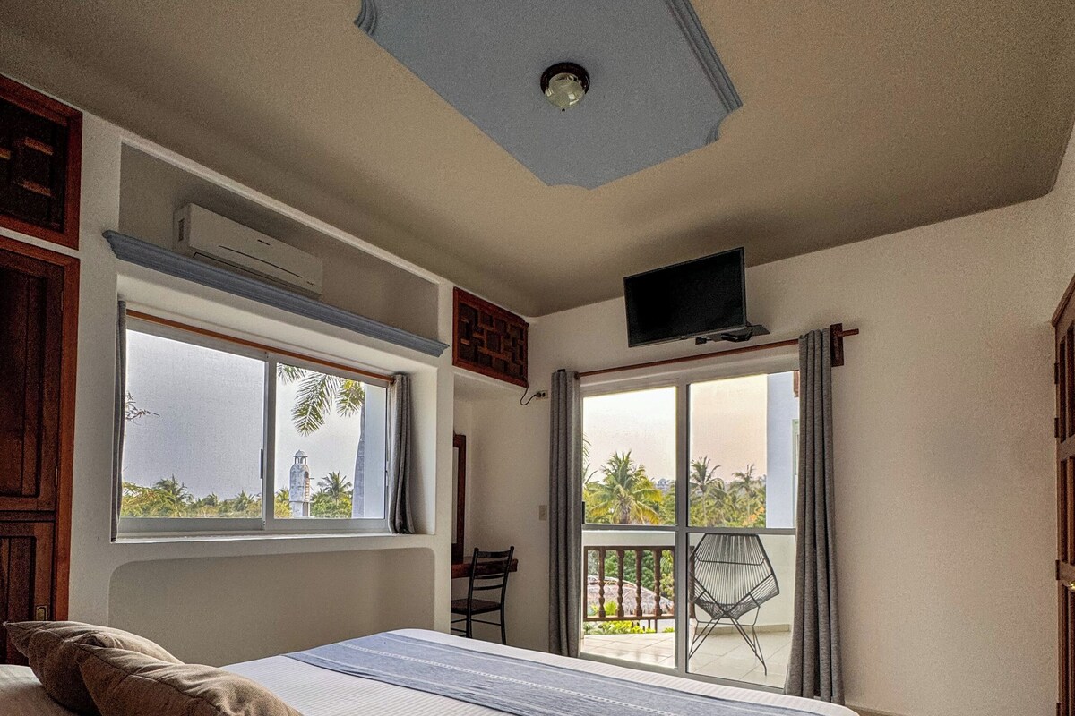 A bedroom with a bed, a television, an air conditioning unit, a window, and a balcony with a view.