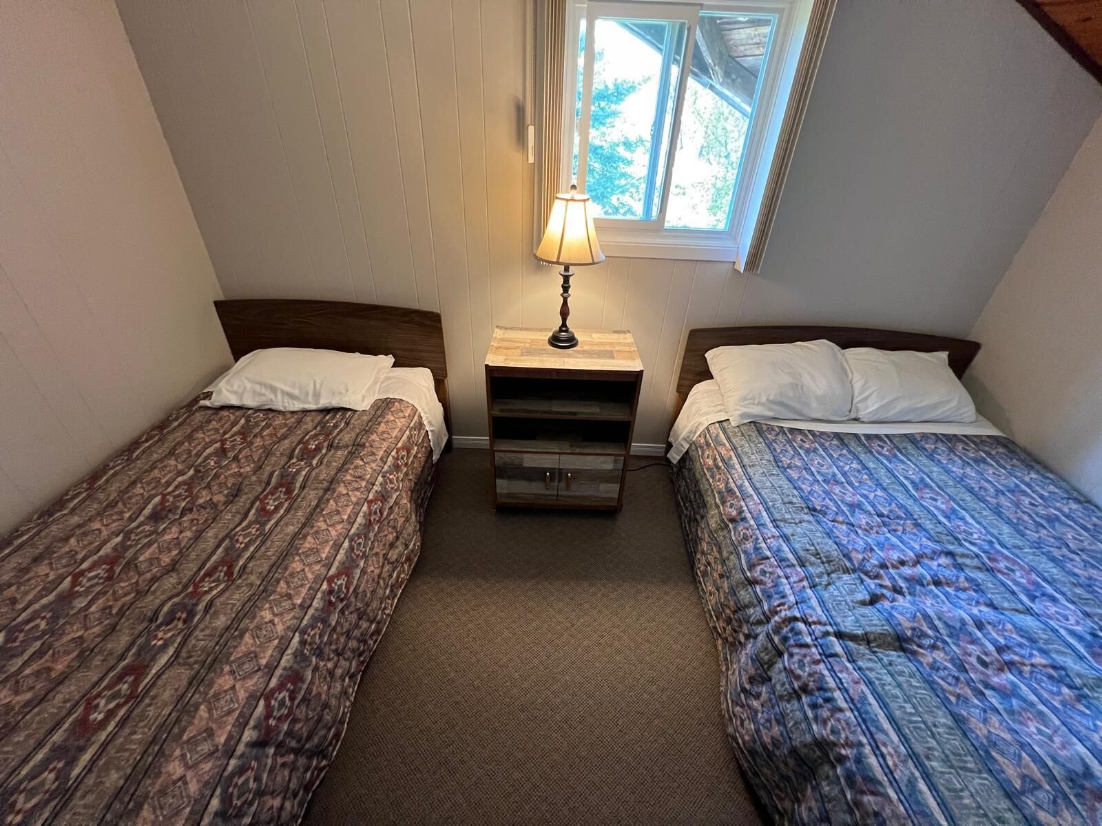 A small, cozy room with two single beds, a nightstand with a lamp, and a window providing natural light.