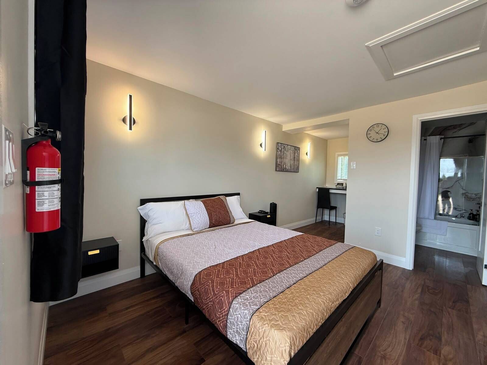A bedroom with a bed, bedside tables, a desk, a chair, a fire extinguisher, and a clock on the wall.