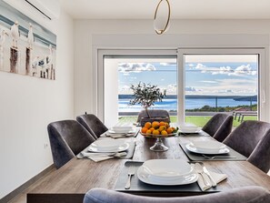 Un comedor con una mesa preparada para cuatro personas, un jarrón de naranjas y vista al mar a través de amplios ventanales.