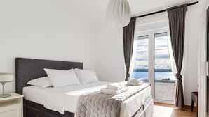 A bedroom with a bed, a nightstand, a bedside table, a window with curtains, and a view of the sky. - Silencia by Interhome (Maslenica)
