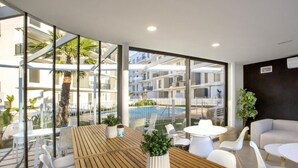 Uma área de estar moderna ao ar livre, com mesa de madeira e cadeiras brancas, com vista para uma piscina e edifícios residenciais.