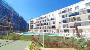 Um edifício de apartamentos moderno com piscina, rodeado por uma cerca branca e vegetação.