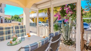 A sunlit patio with a table set for two, surrounded by wicker chairs, a large aloe vera plant, and a view of the street with parked cars and buildings. - Lovely apartment in Vir with WiFi (Vir)