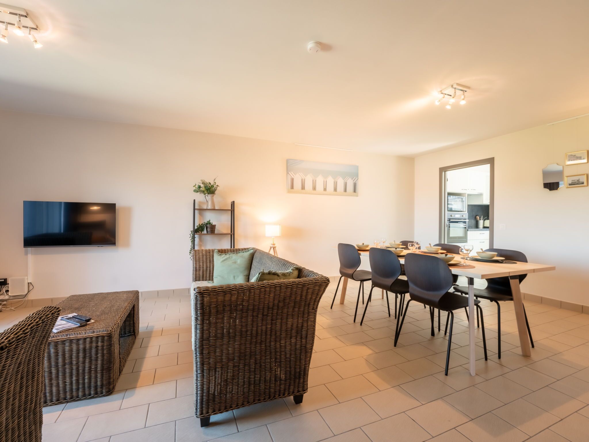 A modern living room with a TV, wicker furniture, a dining area with chairs and a table, and a kitchen visible through an open door.