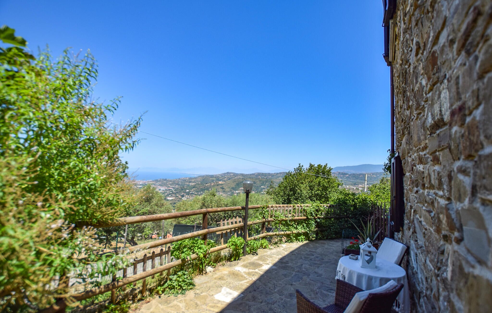 A stone building with a patio area featuring a table and chairs, overlooking a scenic landscape with hills and a clear blue sky.
