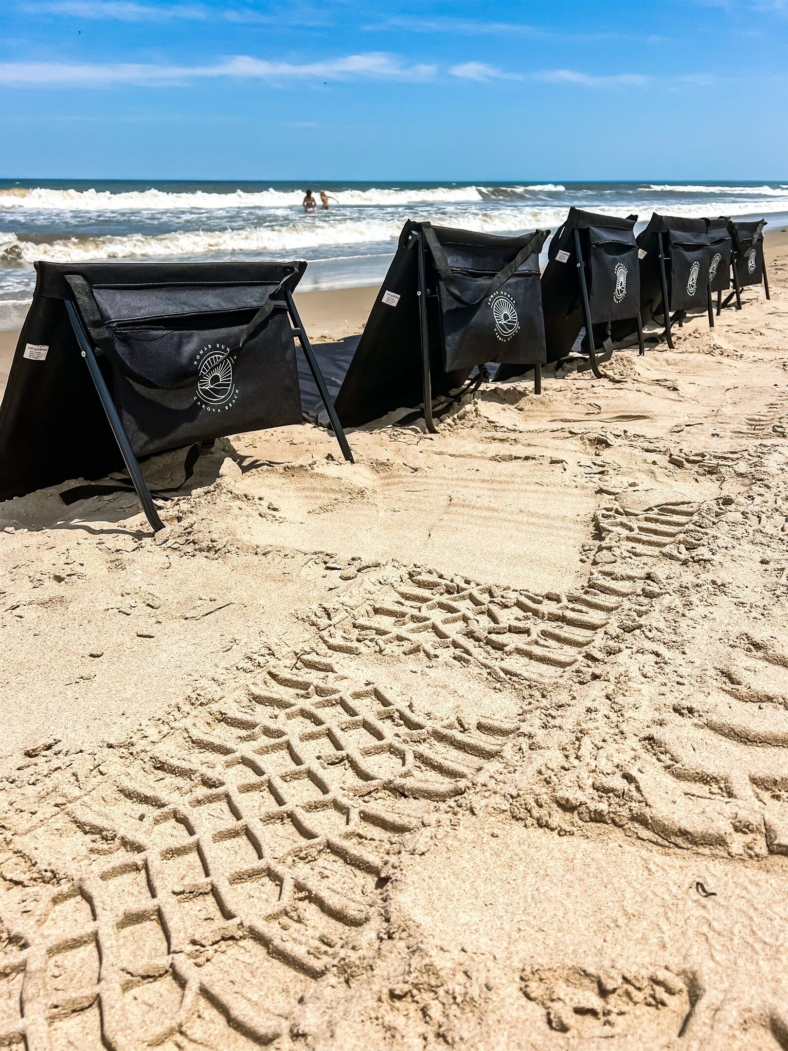 Fileira de cadeiras de praia pretas com logotipos numa praia arenosa.