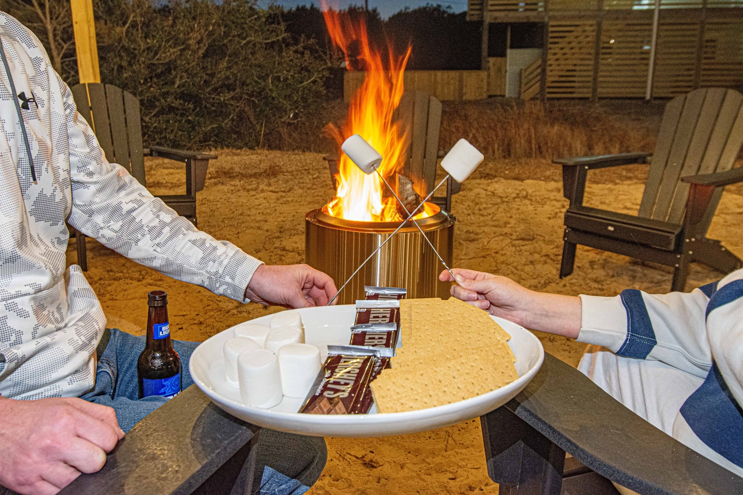 Een kampvuur met marshmallows, chocoladerepen, een biertje en een bord met snacks.