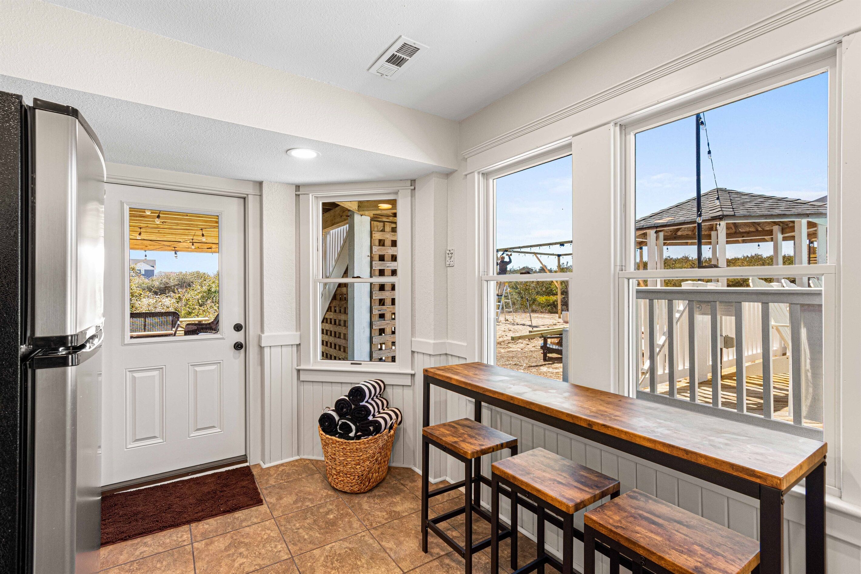 A kitchen with a white door, a wooden bench, a wicker basket with rolled-up mats, and a view of a deck through a large window.