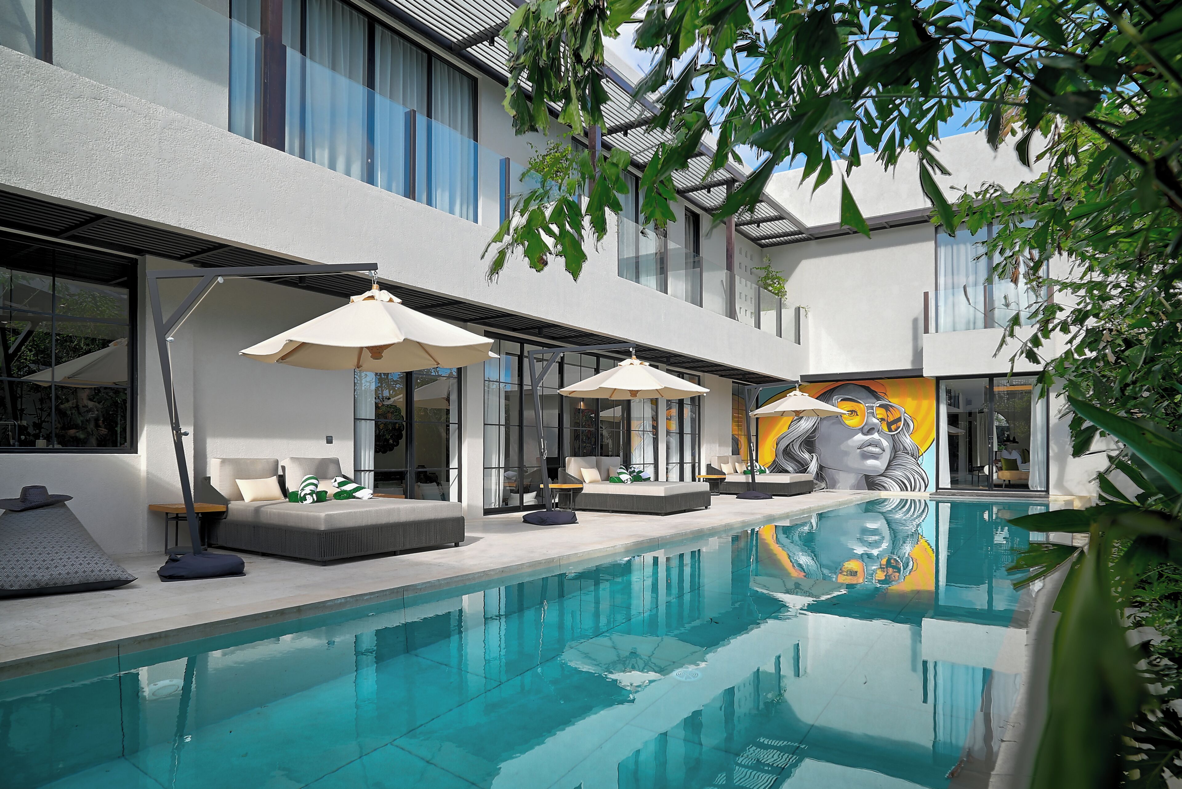 Uma área de piscina moderna ao ar livre, com espreguiçadeiras e guarda-sóis, rodeada por um edifício com grandes janelas e um mural.