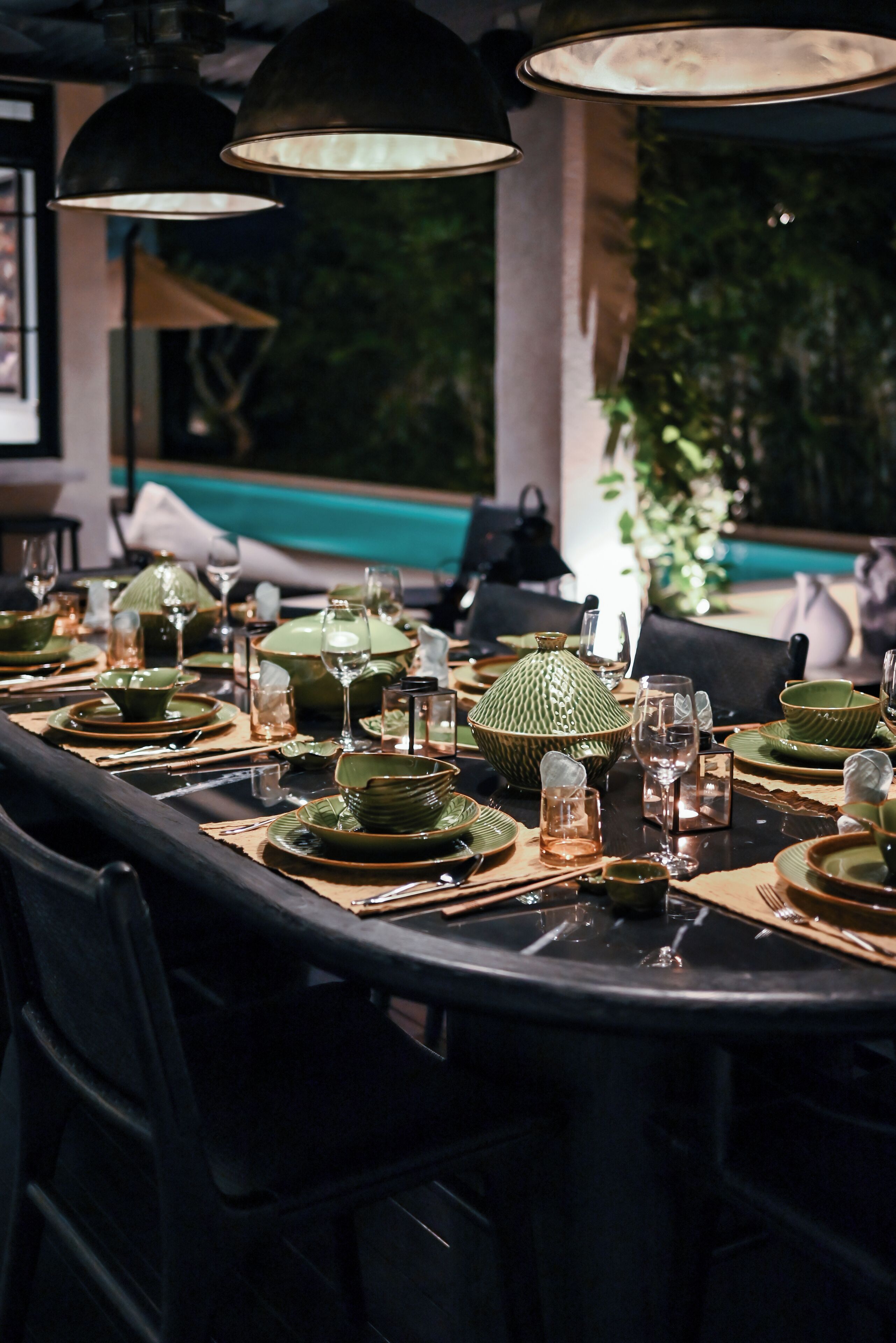 Eettafel gedekt met groene borden, glazen en een grote groene kom, verlicht door zwarte hanglampen.
