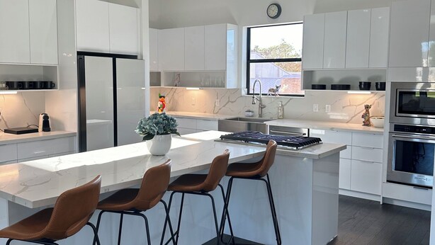 Une cuisine moderne avec des meubles blancs, un îlot central doté de tabourets, un four encastré et une horloge au mur.
