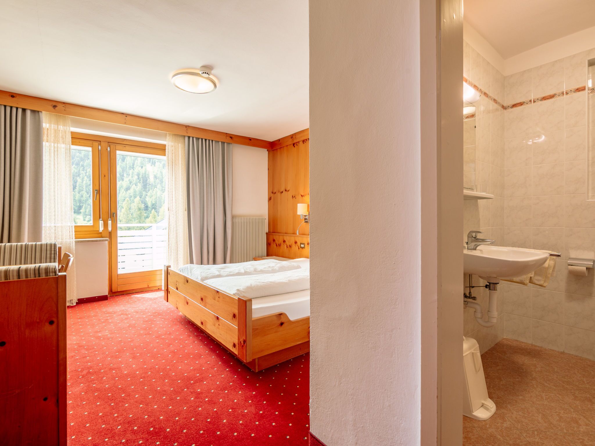 A hotel room with a wooden bed, a red carpet, a window with curtains, and a bathroom with a sink and mirror.