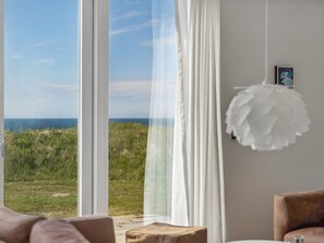 Una habitació amb una finestra gran, una cortina blanca, una lluminària penjant i un quadre amb marc a la paret.