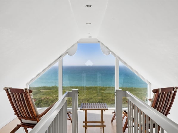 A balcony with a view of the ocean, wooden chairs, and a table.