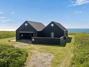 Un bâtiment moderne à deux étages, doté d’un garage, situé sur une colline verdoyante et offrant une vue sur la mer.