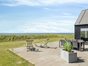 Une terrasse dotée de mobilier d’extérieur, offrant une vue sur la mer, et un bâtiment à l’aspect sombre.