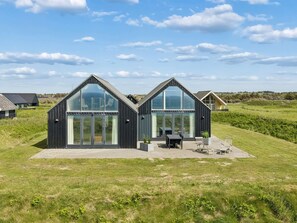 Une maison moderne à deux étages, dotée de grandes fenêtres en verre, d’un patio avec des meubles d’extérieur et d’un jardin verdoyant.