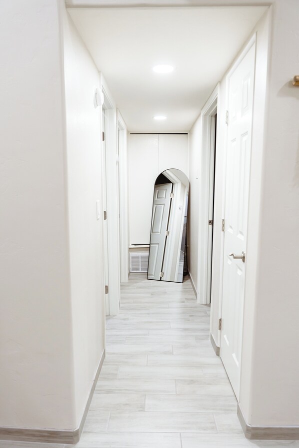 Un pasillo estrecho con paredes blancas y suelo de baldosas, que conduce a una habitación con una puerta al final.