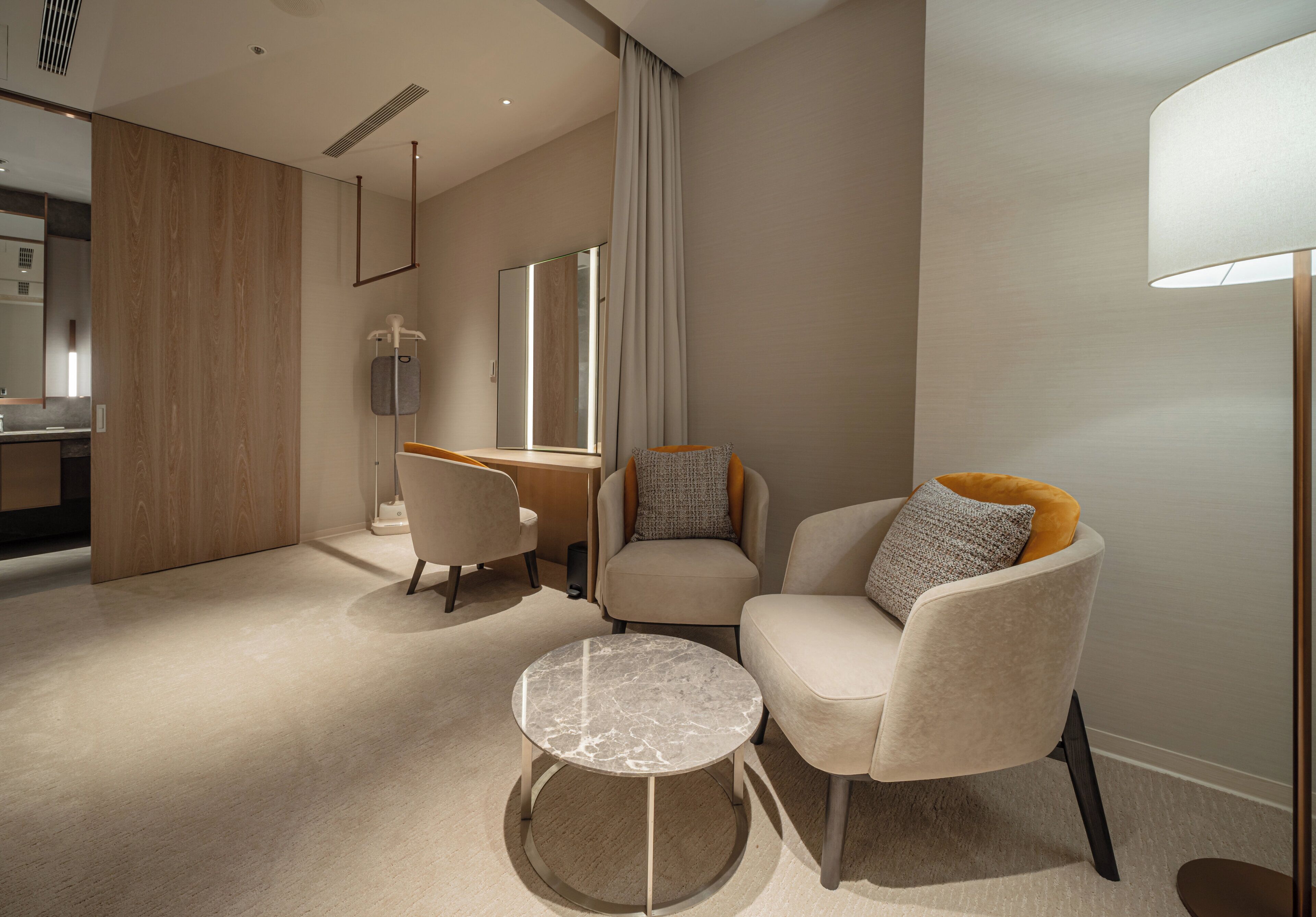 A modern hotel room with a wooden partition, a round marble table, two armchairs, a sofa, a desk, and a large window with curtains.