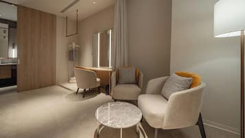 A modern hotel room with a wooden partition, a round marble table, two armchairs, a sofa, a desk, and a large window with curtains.