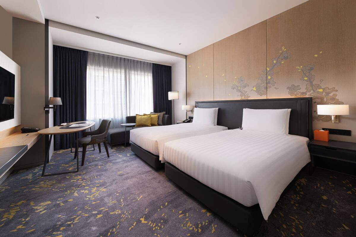 A hotel room with two beds, a desk, a chair, a small table, and a large window with curtains.