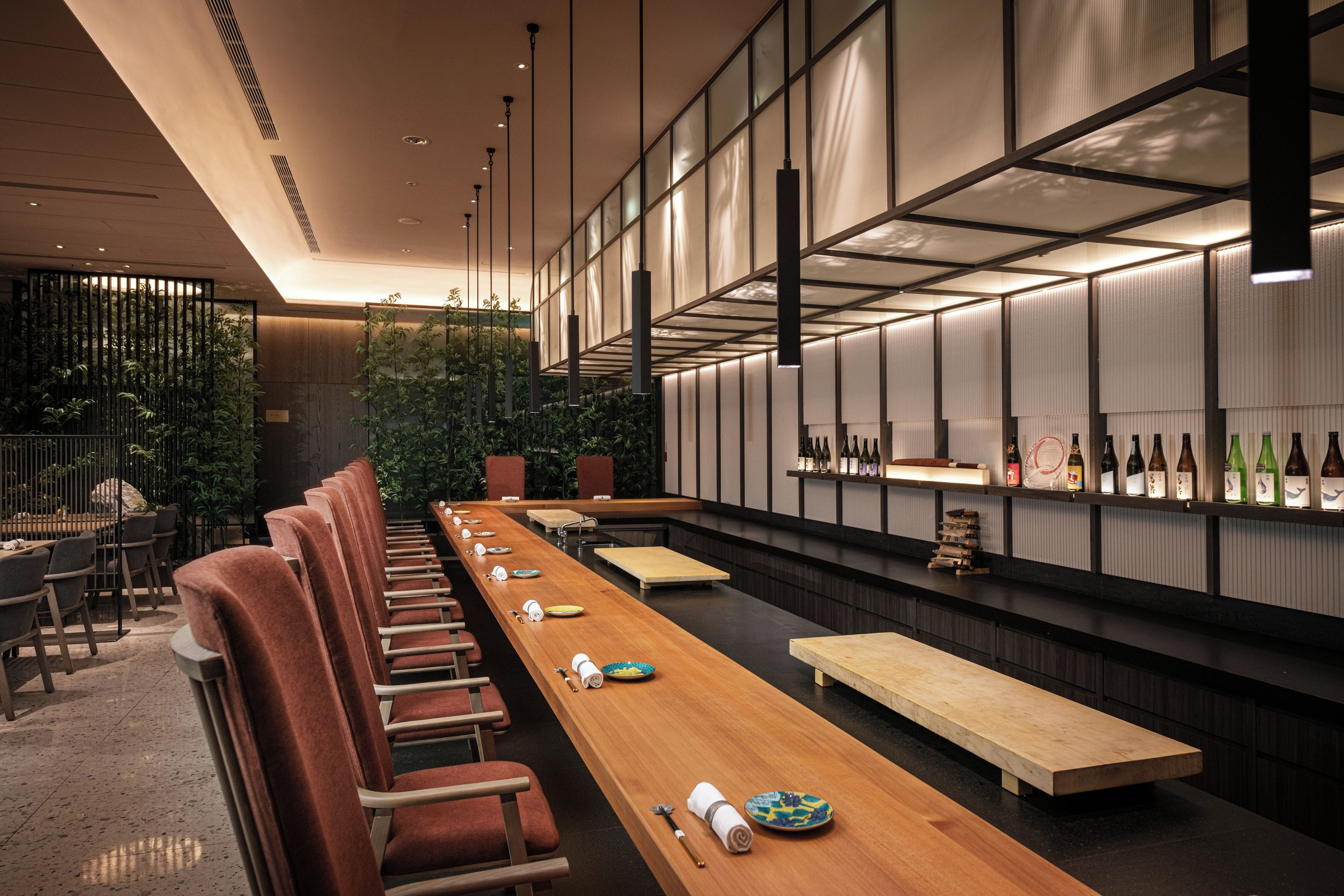 A modern restaurant interior with a long wooden table, cushioned chairs, and a wall of wine bottles.