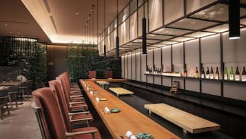 A modern restaurant interior with a long wooden table, cushioned chairs, and a wall of wine bottles.